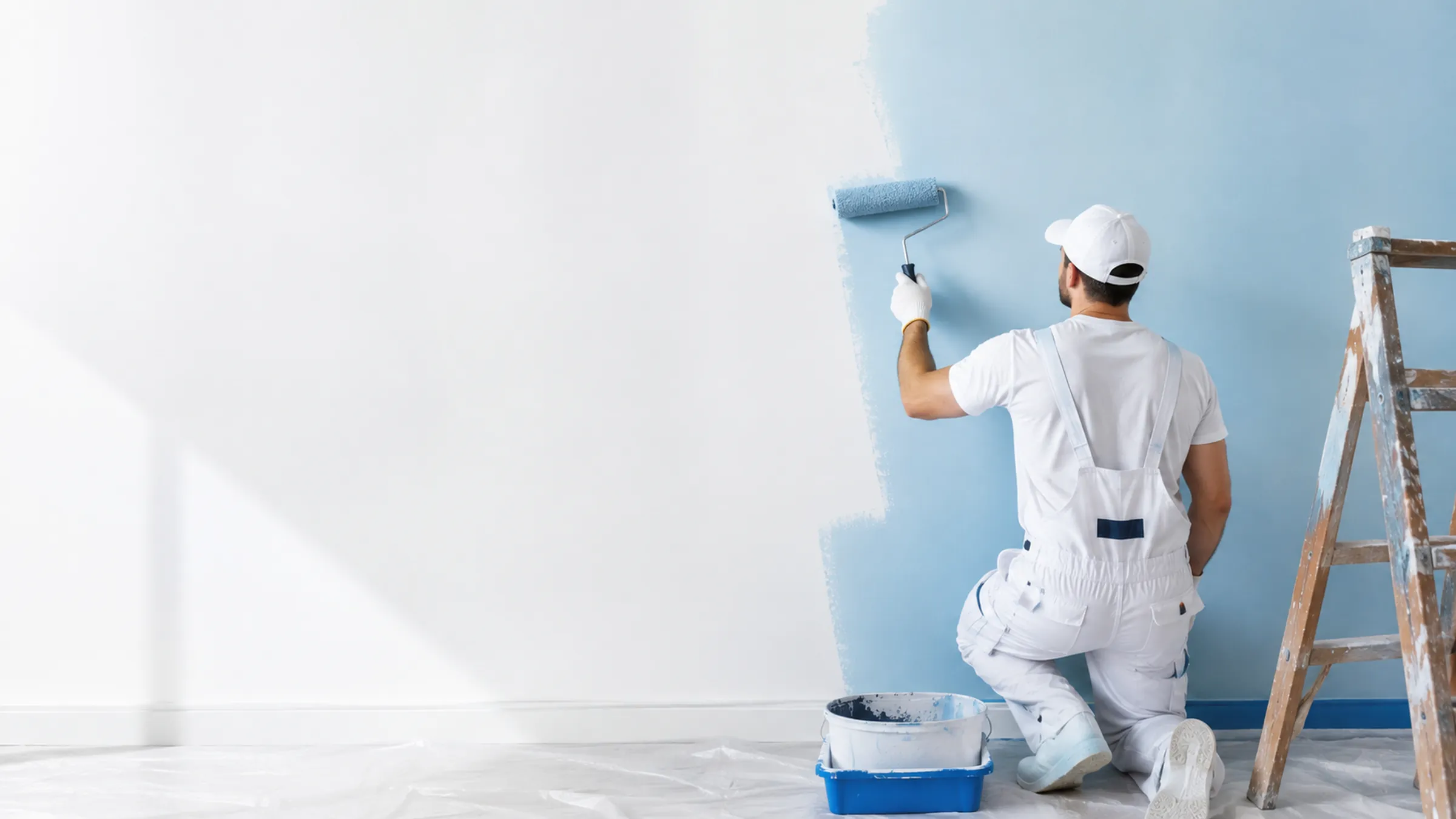 Maler in weißer Arbeitskleidung rollt eine Wand an, daneben Farbeimer und Leiter